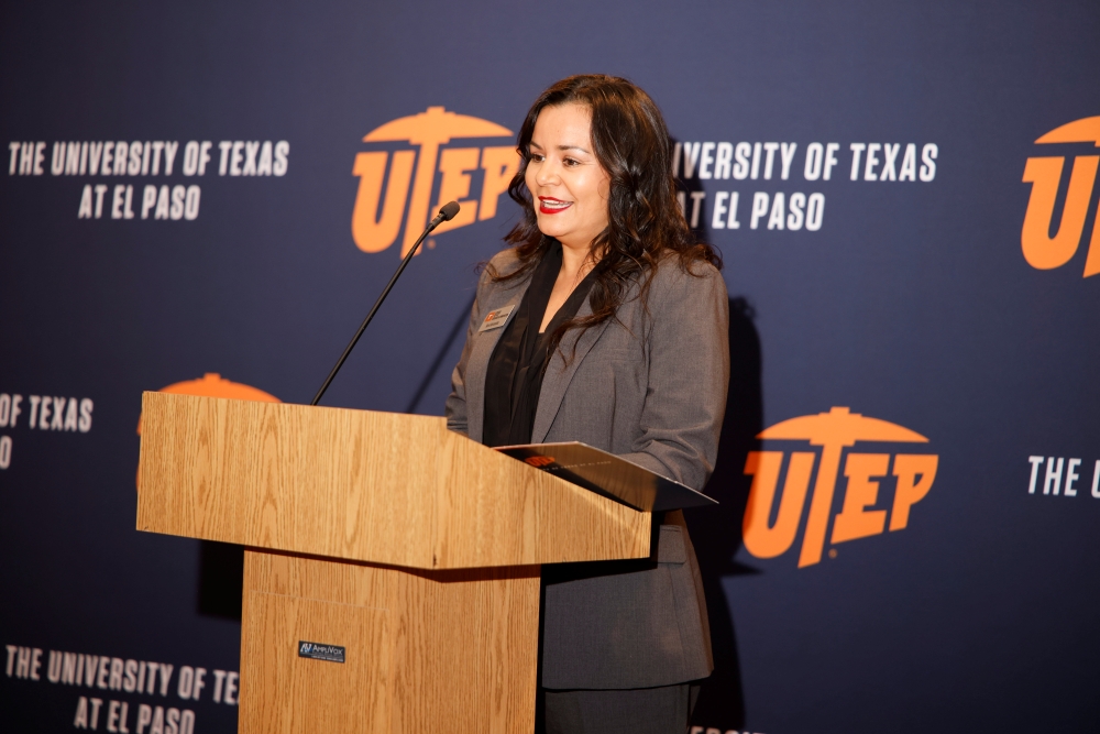 A woman in a gray suit delivers remarks from a podium at a UTEP event.